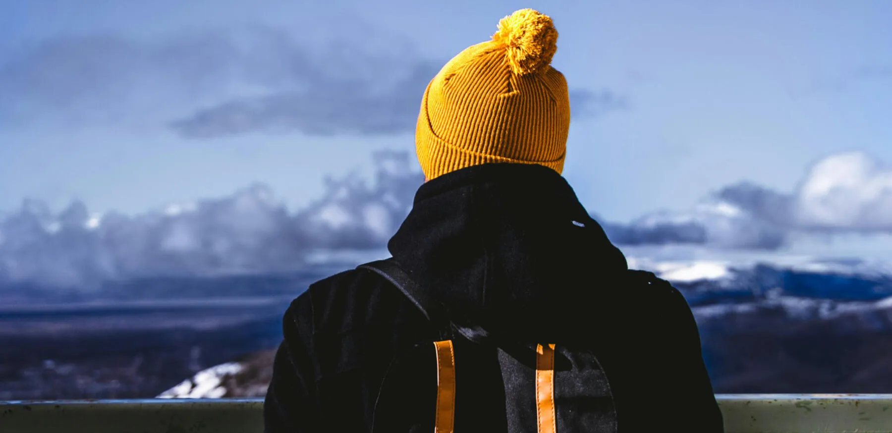 Une personne de dos, portant un bonnet jaune, face à un paysage enneigé.